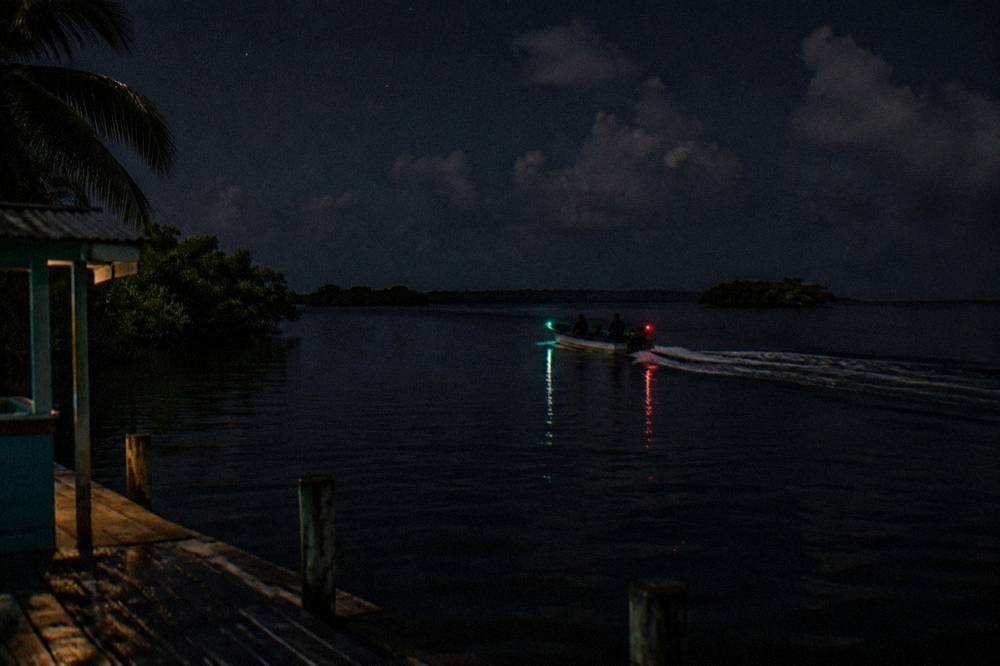 A small boat with navigation lights crossing dark calm water toward distant mangroves