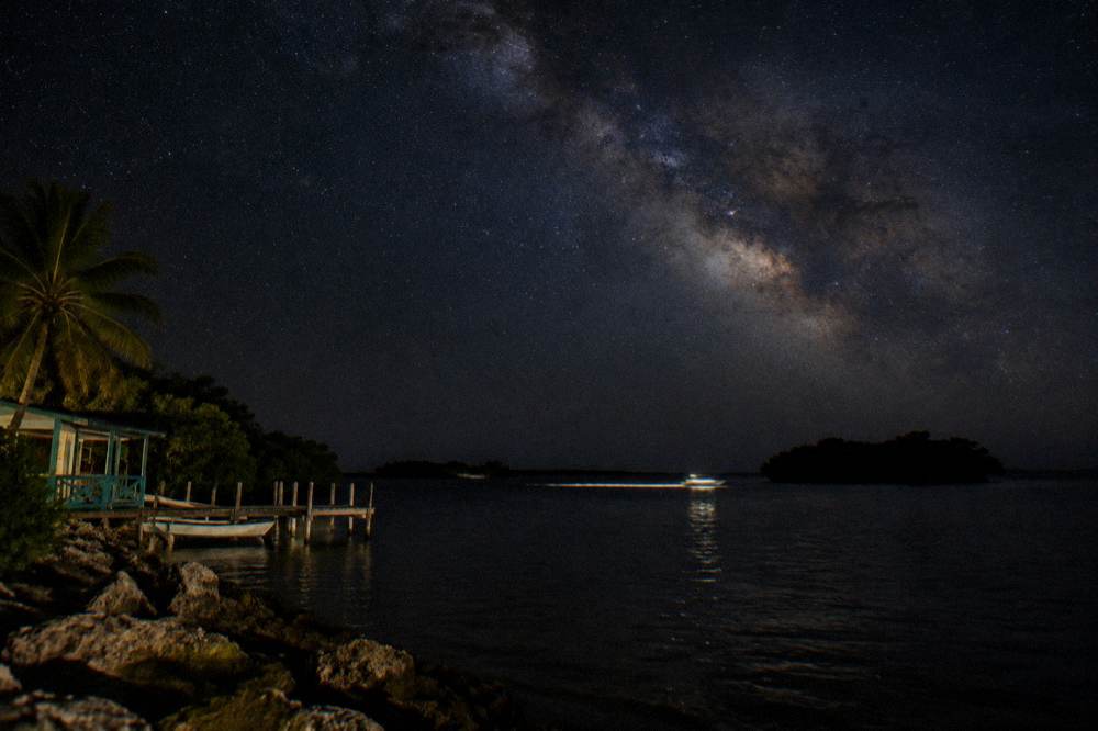 Dark nighttime bay with a line of pale light trailing in the water and stars overhead