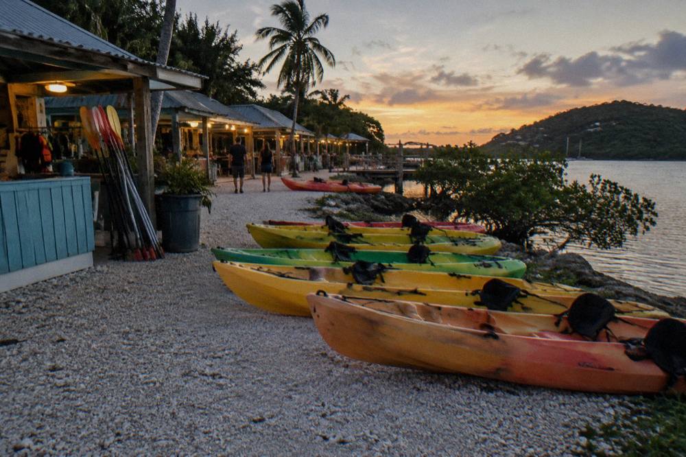 A line of tandem kayaks resting on a dark shoreline beside a small village of kiosks at dusk