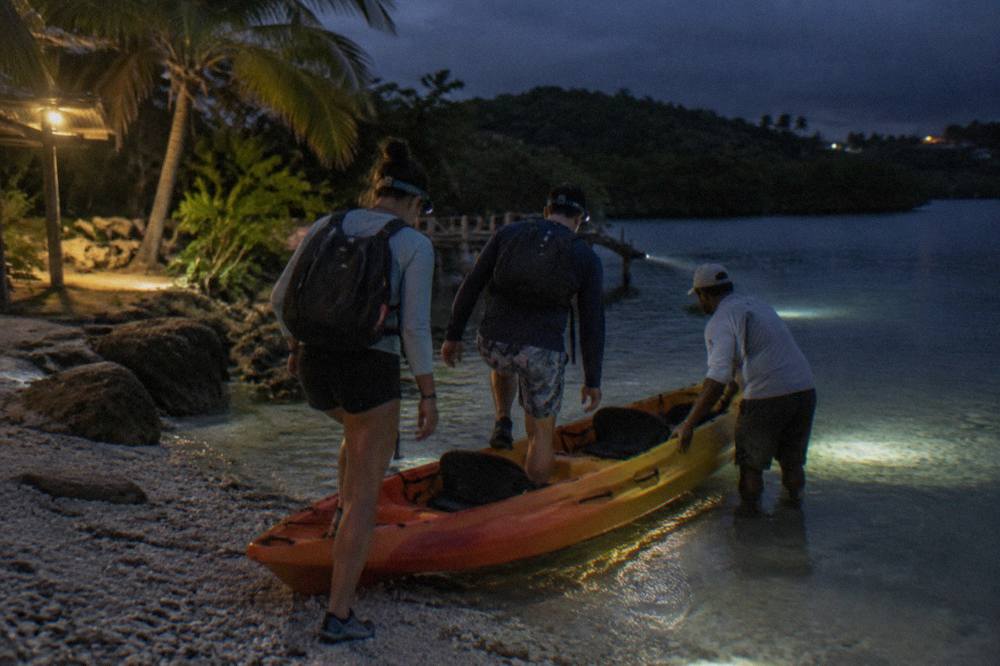 A guide checking a tandem kayak and paddles by headlamp at a dark launch area before a night tour