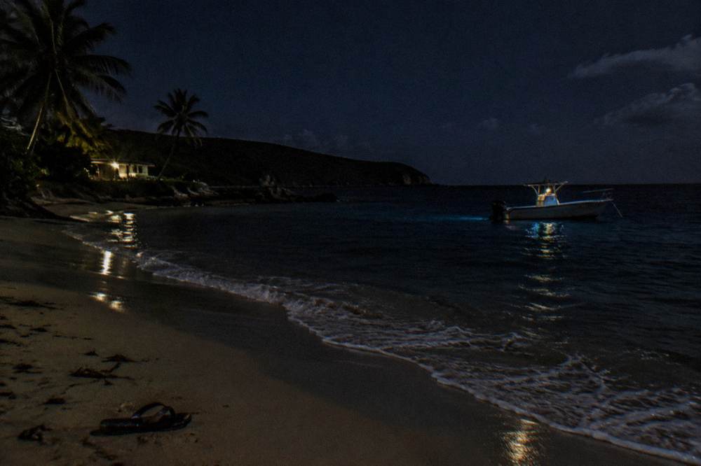 A small boat anchored on dark water at night with faint blue ripples spreading from the hull