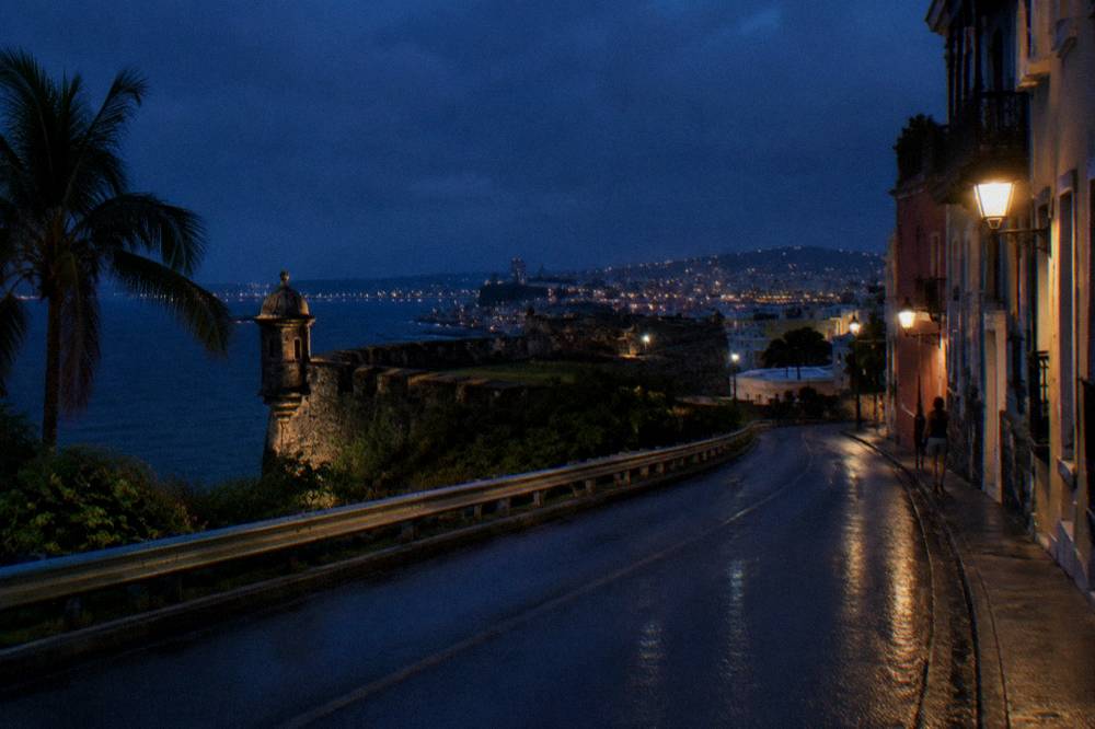 A dark highway stretching east through hills with a town's lights in the distance