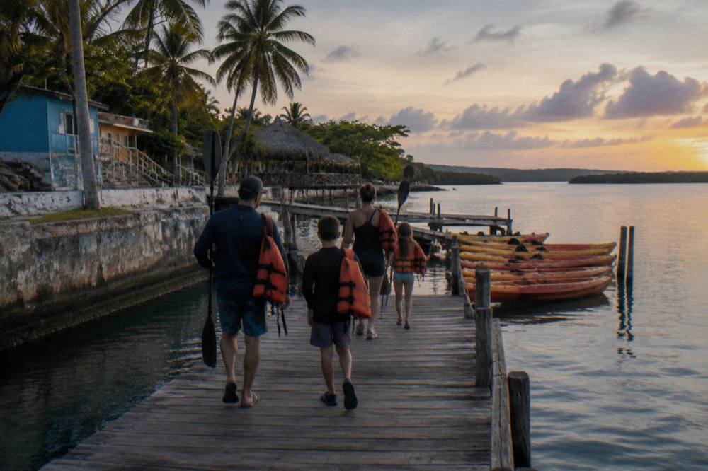 A family of four carrying paddles and life jackets toward kayaks at a dock at dusk