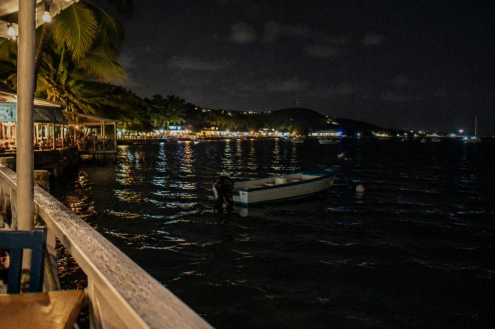 Small boat at anchor on open dark bay water with lights from a distant shoreline