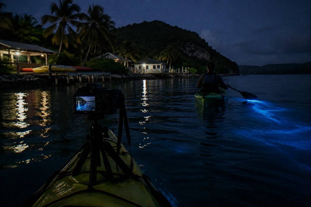 A camera on a tripod at the edge of a dark lagoon, pointed at a kayak with a faint blue glow trailing behind it.
